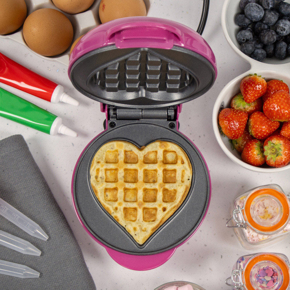 Pink and black heart-shaped waffle maker with golden cooked waffle, surrounded by fresh strawberries, blueberries, eggs, and baking supplies on white surface