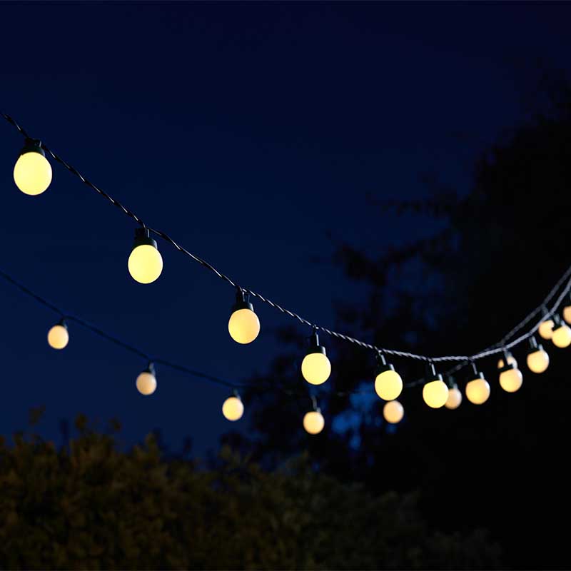 String of 30 warm yellow spherical LED festoon lights draped against a deep blue evening sky with silhouetted trees in the background