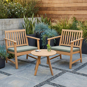 Kaemingk Acacia wood 2 seater garden bistro set with two natural wooden chairs with vertical slat backs, grey cushions, and a round matching wooden table on a paved patio with decorative border, surrounded by potted plants and wooden fence