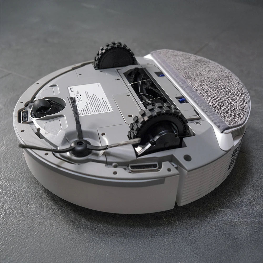 White circular robotic vacuum cleaner with black dual rotating brushes, gray fabric dust collection pad on top, control panel with documentation, power cord, and tread wheels visible from overhead angle on gray surface