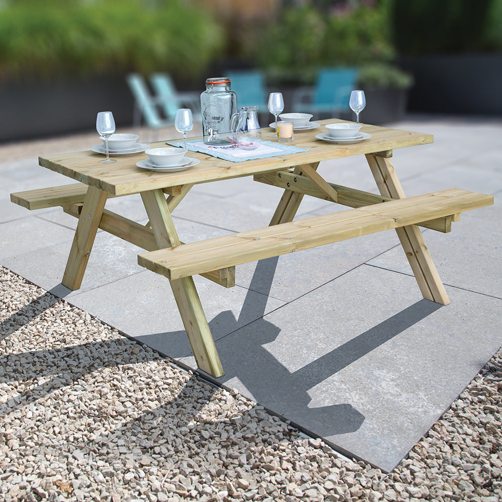 Pressure treated wooden picnic bench table with attached benches set outdoors on patio, laid with white dinnerware, wine glasses, and table linens in a garden setting