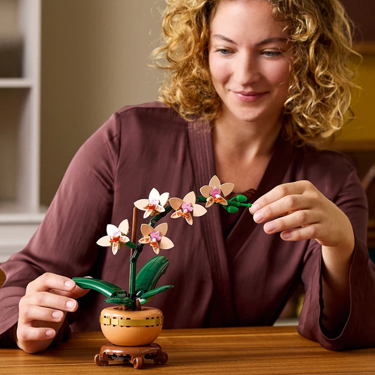 Woman with curly auburn hair wearing a burgundy shirt admiring a LEGO Orchid set featuring white and pink flowers with green leaves in a wooden pot on a wooden table