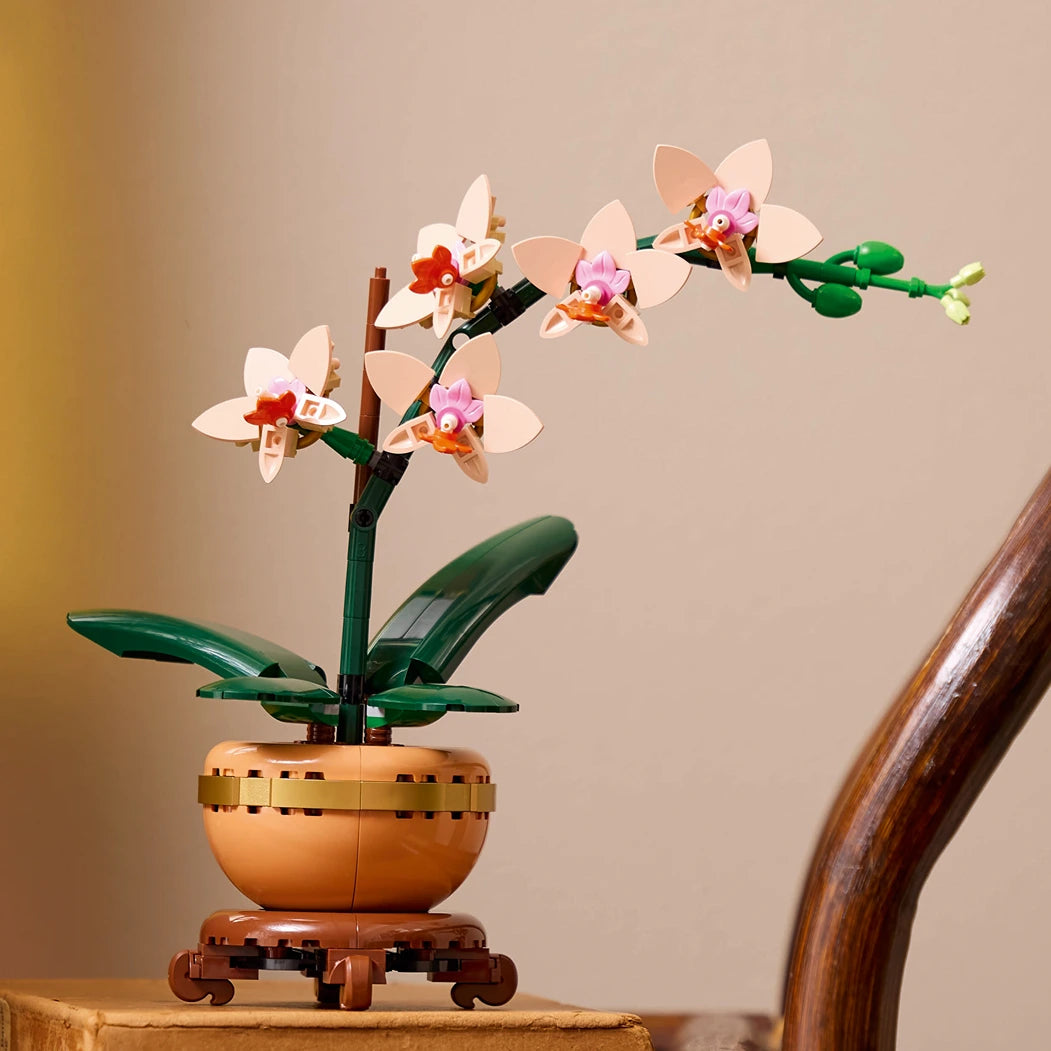 LEGO Orchid building set featuring white and pink flowers with orange centers on green stems in a terracotta pot with gold trim, displayed on wooden surface against neutral background