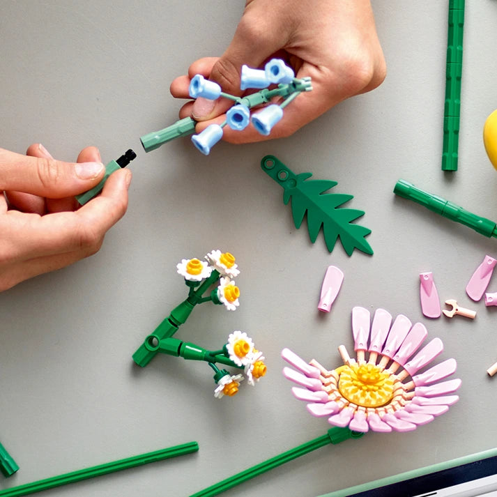 Child's hands assembling LEGO Petite Sunny Bouquet set with white and yellow daisy flowers, green stems and leaves on gray surface, showing completed pink gerbera and pea pod designs