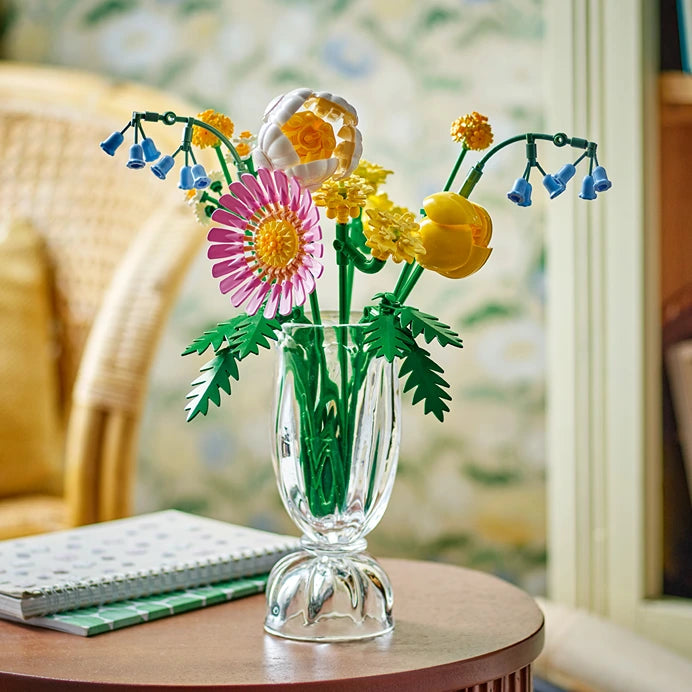 LEGO Petite Sunny Bouquet set 10347 featuring colorful buildable flowers including pink gerbera, yellow roses, and blue bellflowers with green stems arranged in a clear glass vase on a wooden side table