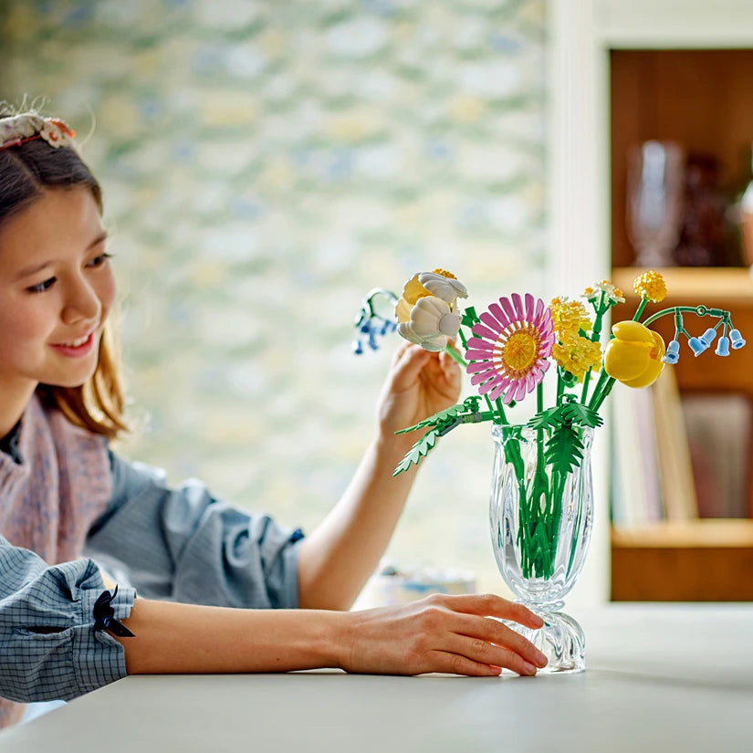Young girl smiling while arranging colorful LEGO flowers including pink gerbera daisies, yellow daffodils, and white blossoms in a clear glass vase with green stems on a white table in a bright room