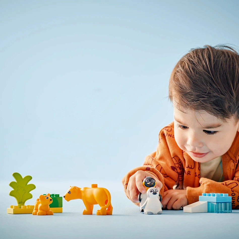 Young child in orange jacket playing with colorful LEGO Wild Animal Families set featuring orange lion and giraffe figurines, green plant piece, and penguin toy against light blue background