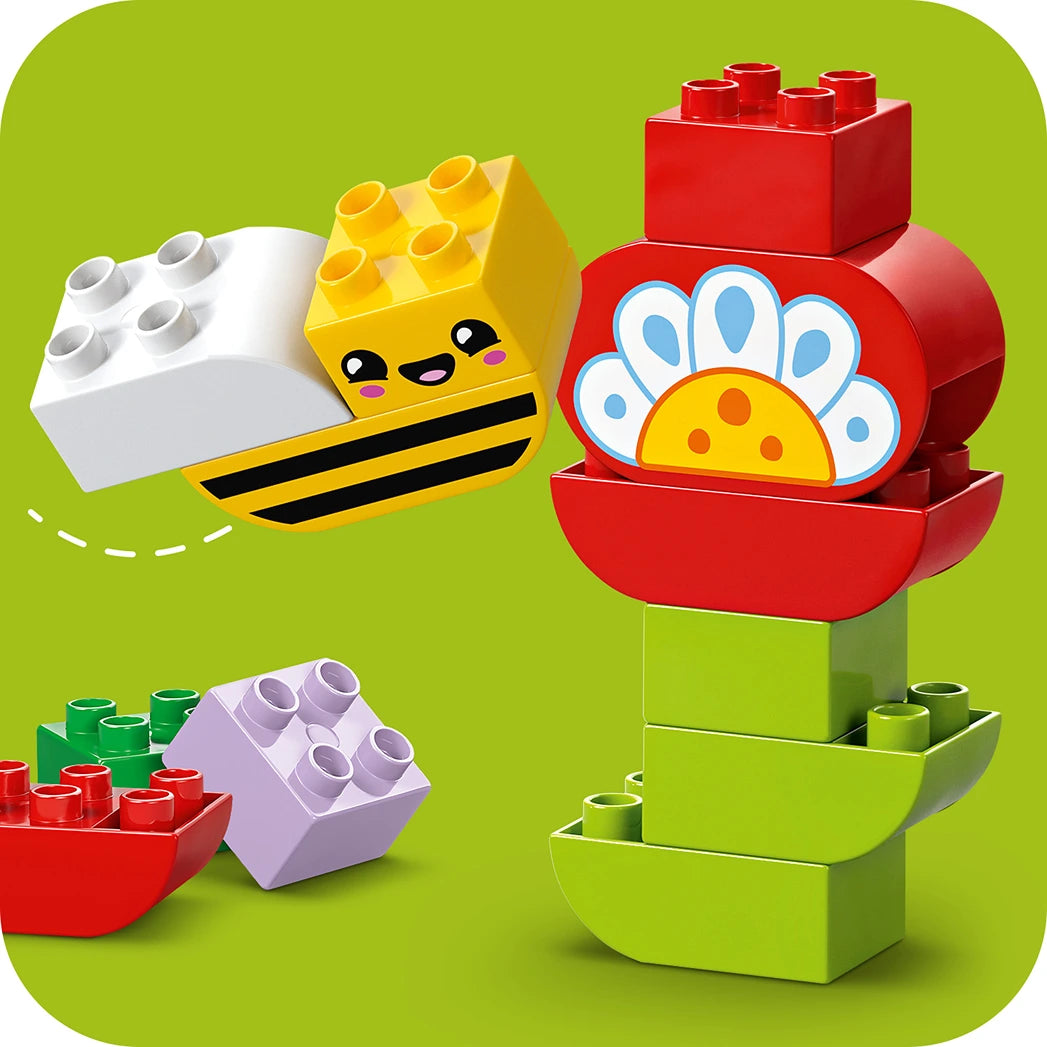 LEGO Creative Garden and Flowers set 10444 featuring a smiling yellow bee brick with white and yellow blocks, red flower with white petals and yellow center, green stem blocks, and assorted red, purple, and green LEGO bricks on a bright green background