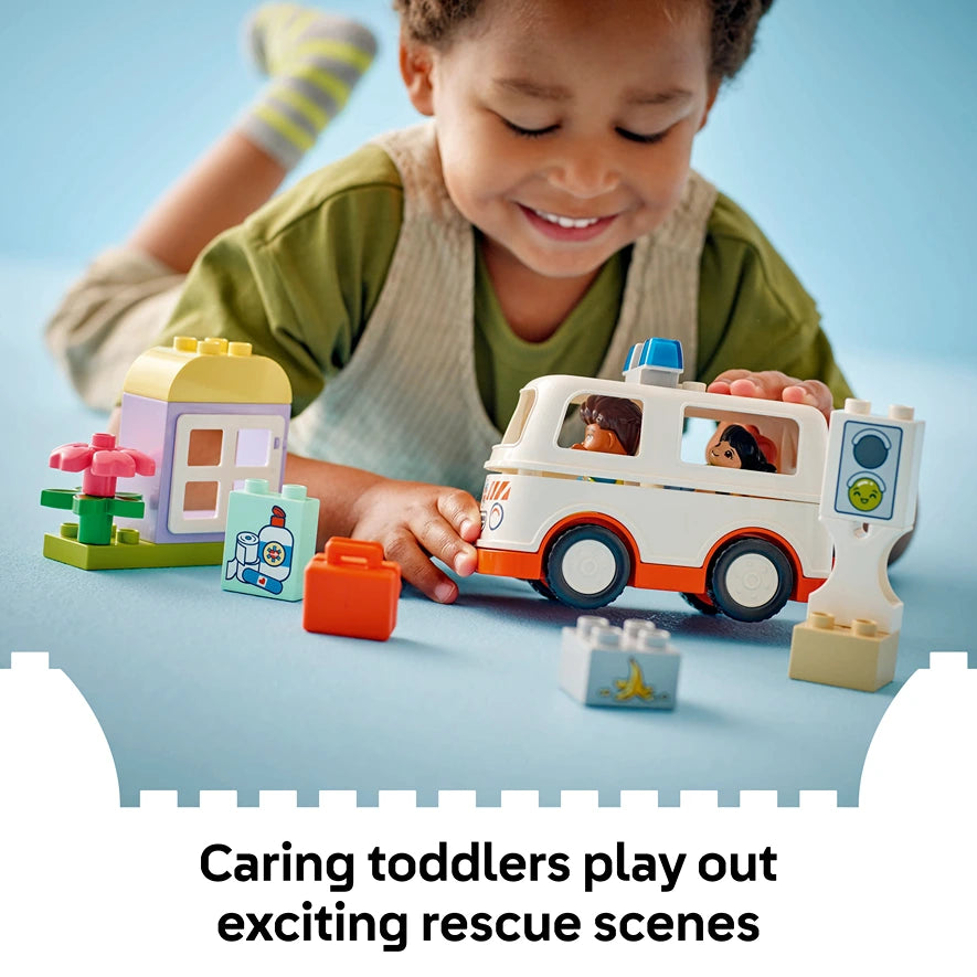 Young child playing with colorful LEGO Duplo ambulance set featuring white and orange ambulance vehicle with blue siren, small figurines, building blocks in green yellow and pink, and accessories on blue surface against light blue background
