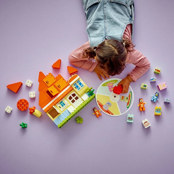 Overhead view of a child playing with Lego Bluey's Family House set featuring a colorful orange and yellow house structure, minifigures, and accessories scattered on a purple background alongside a circular memory game component
