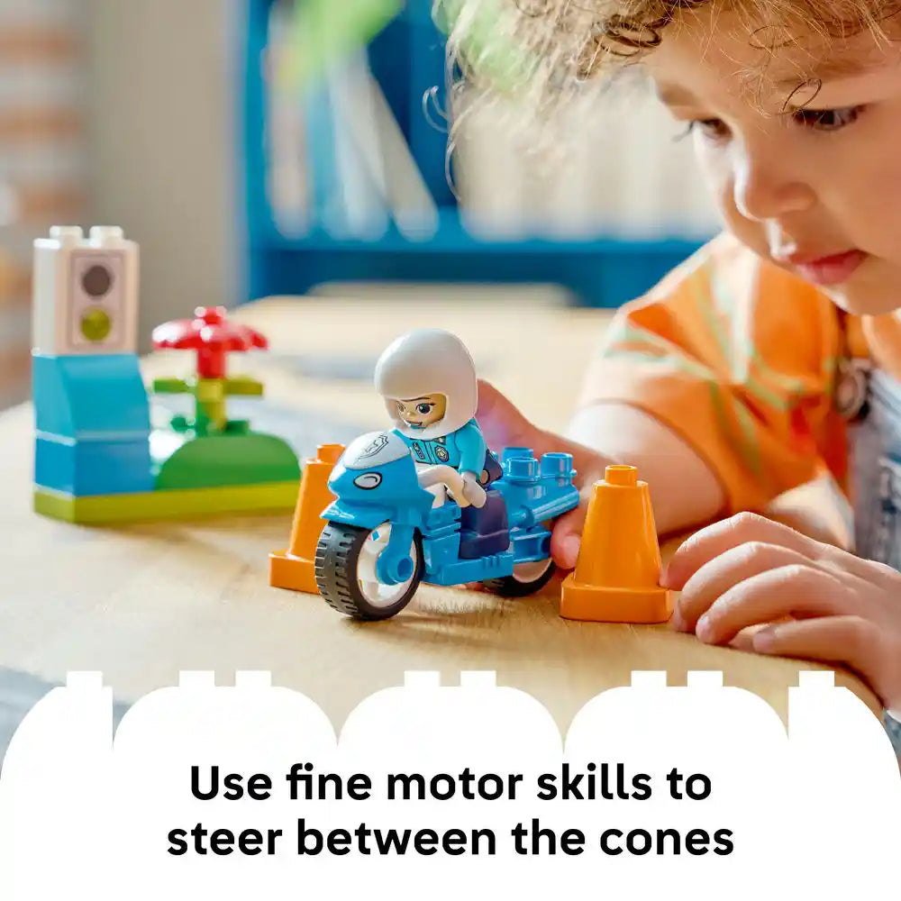 Young child with curly hair wearing orange shirt playing with blue LEGO police motorcycle set, maneuvering toy between orange traffic cones on wooden surface, demonstrating fine motor skills development with colorful building blocks and minifigure in background