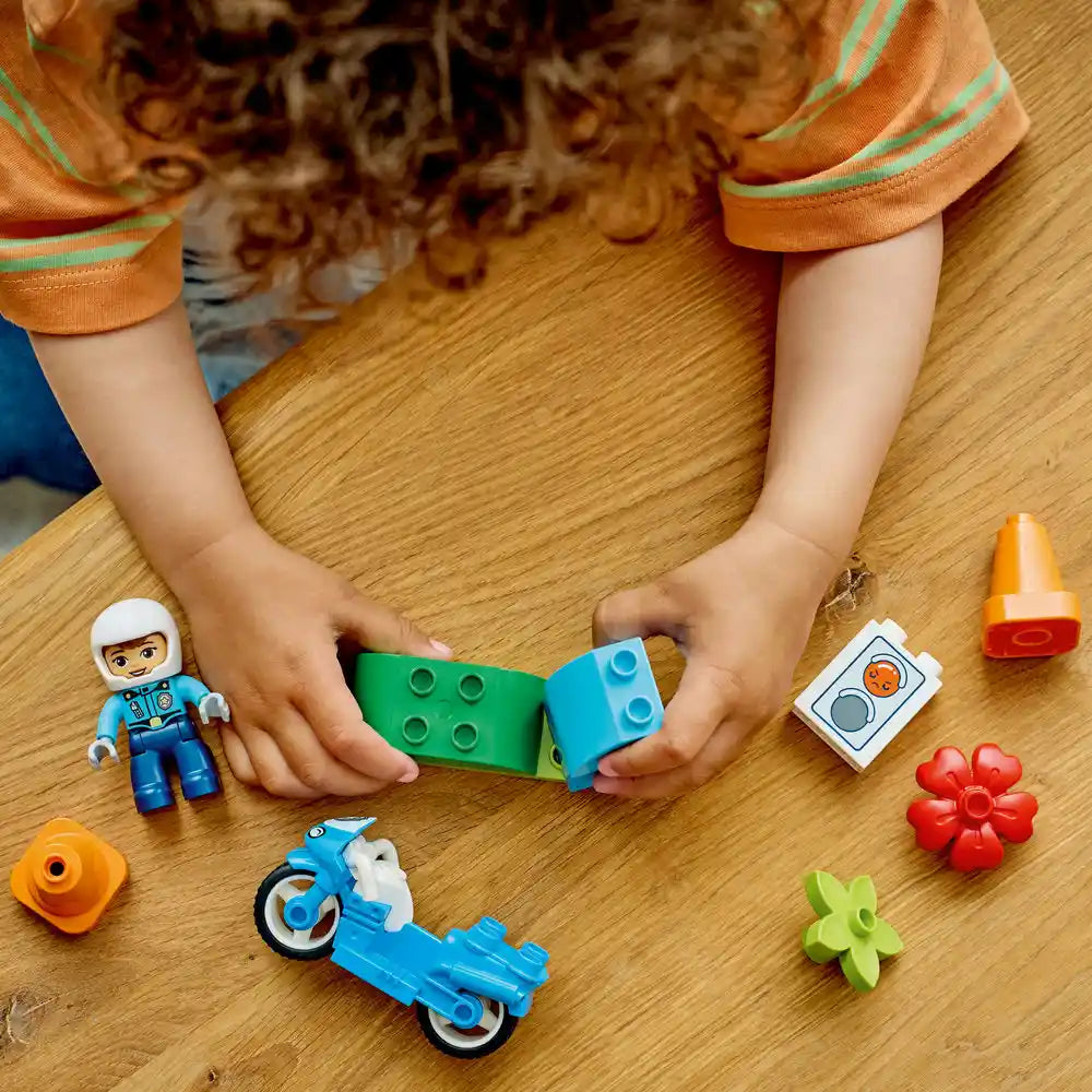 Child's hands holding green and blue LEGO bricks above wooden surface with LEGO Duplo police officer minifigure, blue motorcycle, red flower, orange pieces, and instruction card