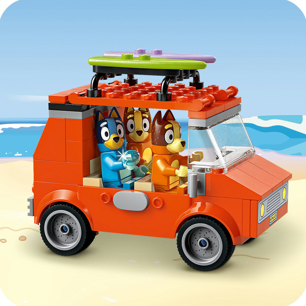LEGO Bluey's Beach & Family Car Trip building set featuring a bright orange brick vehicle with black wheels on sandy beach background, with cartoon dog characters visible in the cabin and a green and purple surfboard on the roof rack against a blue sky and ocean backdrop