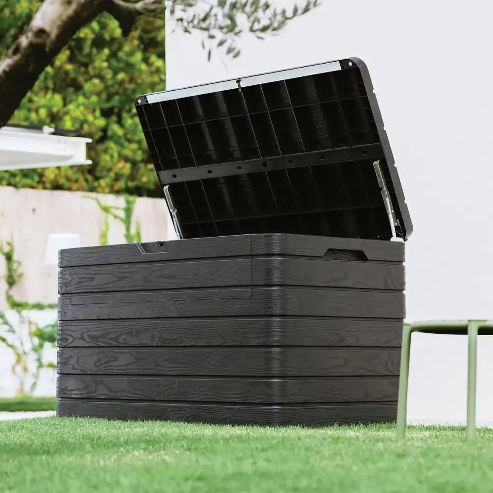 Toomax Dolomiti anthracite garden storage box with open lid showing black interior compartments, positioned on grass in a garden setting with white fencing and green foliage in background