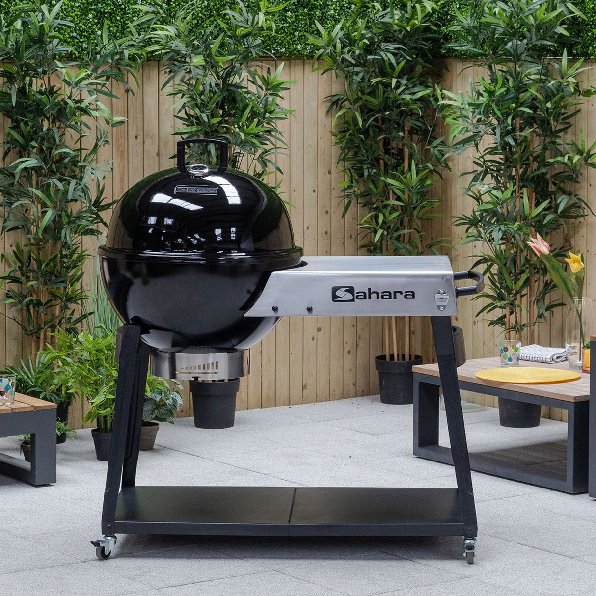 Sahara Forge Host charcoal BBQ grill with glossy black dome lid and white control panel on black wheeled cart in garden patio setting with wooden fence and green plants