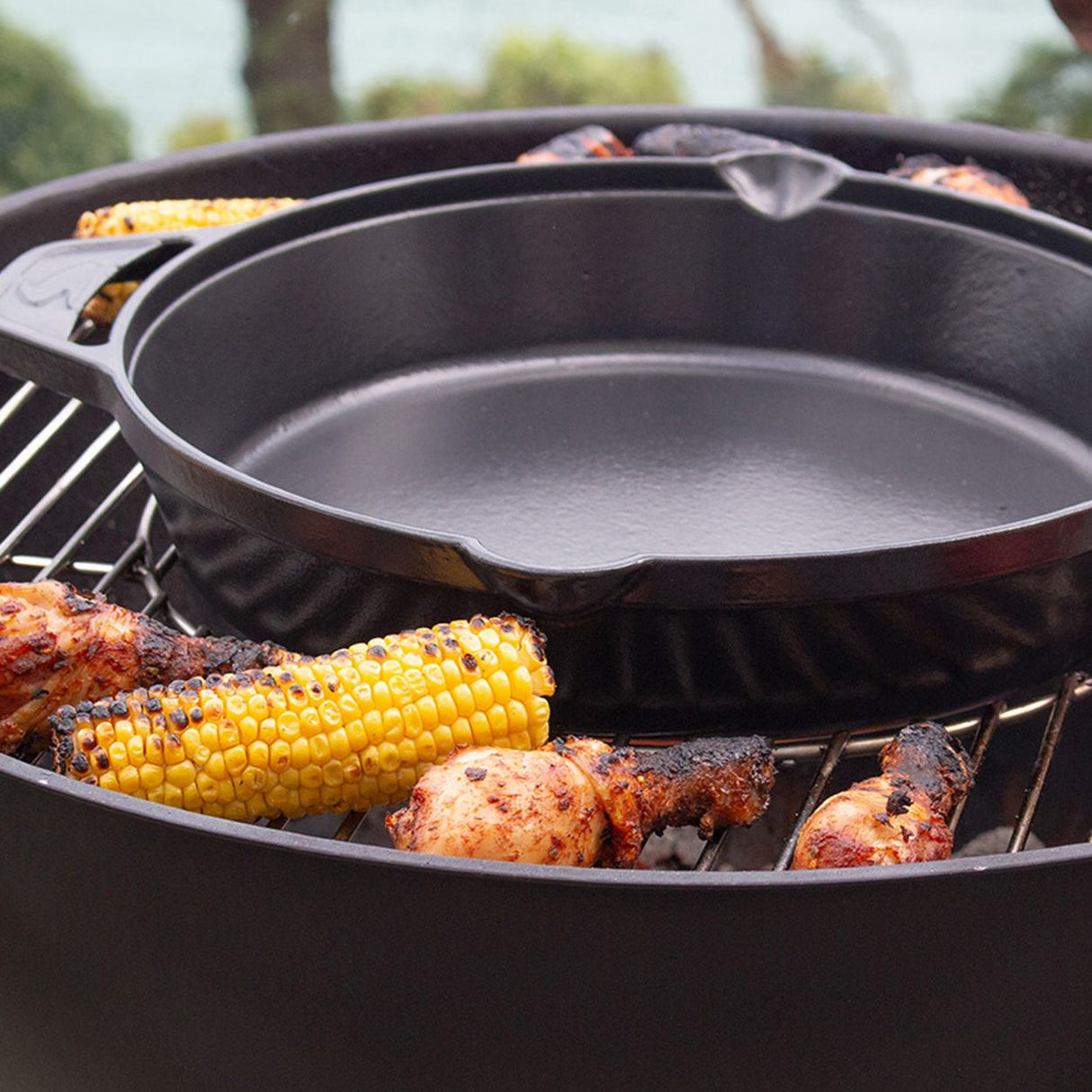Black cast iron charcoal BBQ grill with corn and seasoned chicken cooking on grates, featuring empty griddle pan on top, outdoor garden setting