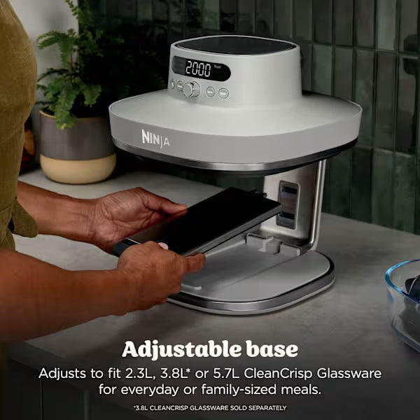 Ninja CRISPi PRO XL air fryer with white dome-shaped body and black base on adjustable stand, displaying 2800 temperature setting, with person's hands adjusting the base to show compatibility with 2.3L, 3.8L or 5.7L CleanCrisp glassware bowls, potted plant visible in background