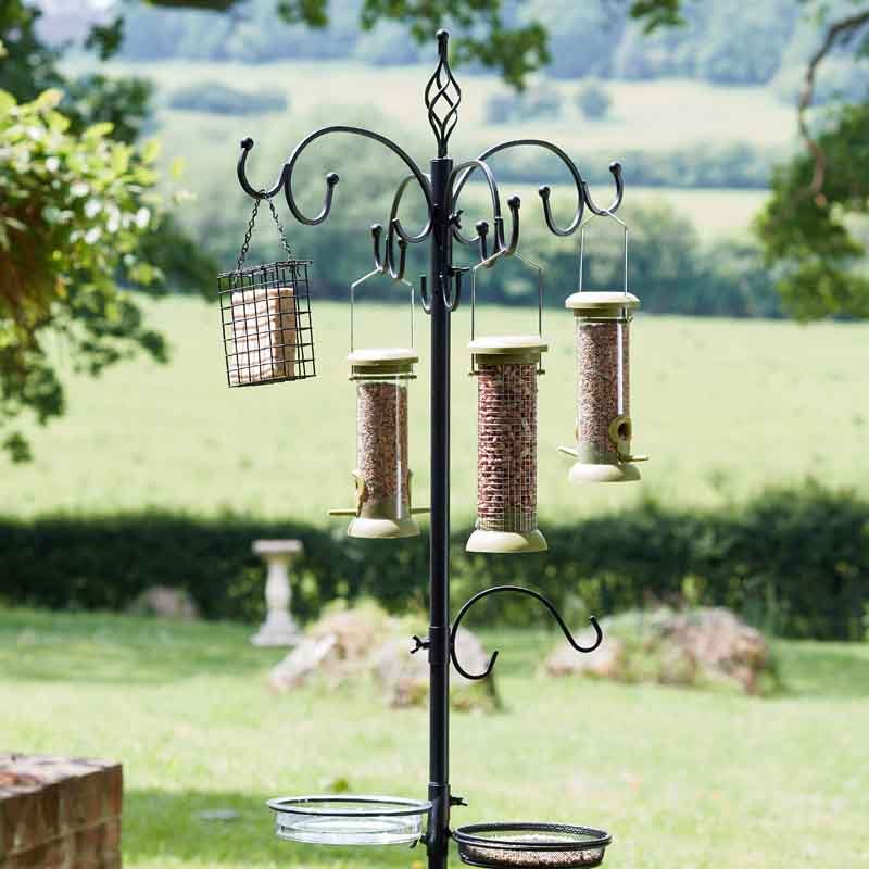 Black wrought iron bird feeding station with multiple hanging seed feeders, suet cage and water dishes in a garden setting with countryside landscape in background