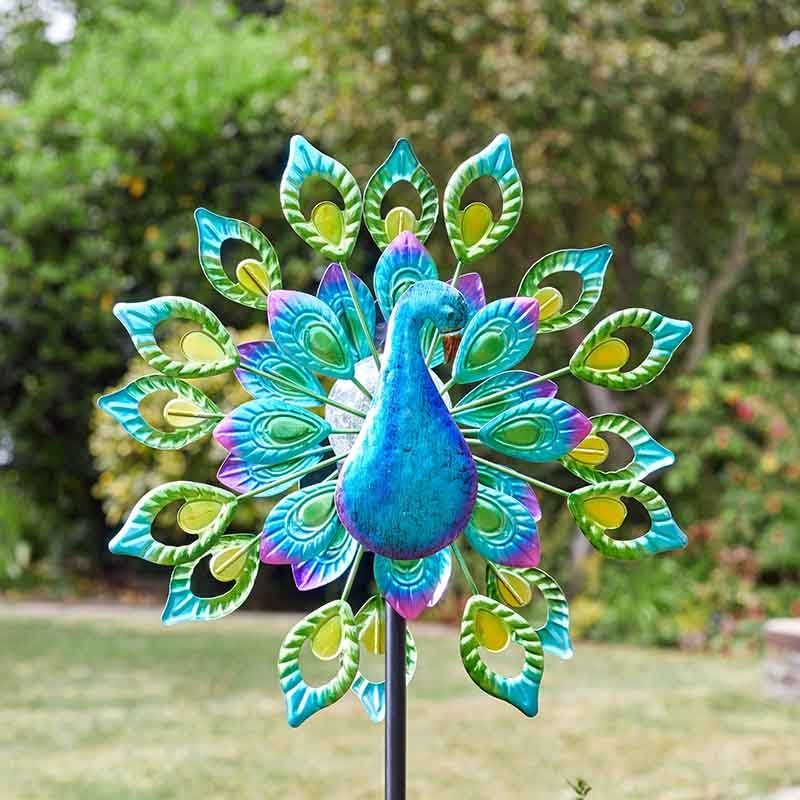 Colorful metal peacock solar wind spinner with turquoise blue body, green and yellow feather details, and purple accents on black pole in garden setting