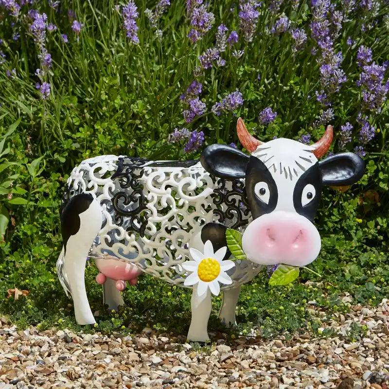 Decorative garden silhouette of a whimsical black and white cow with ornate patterned body, pink nose, brown horns and a white daisy flower, positioned on gravel in front of blooming purple lavender flowers