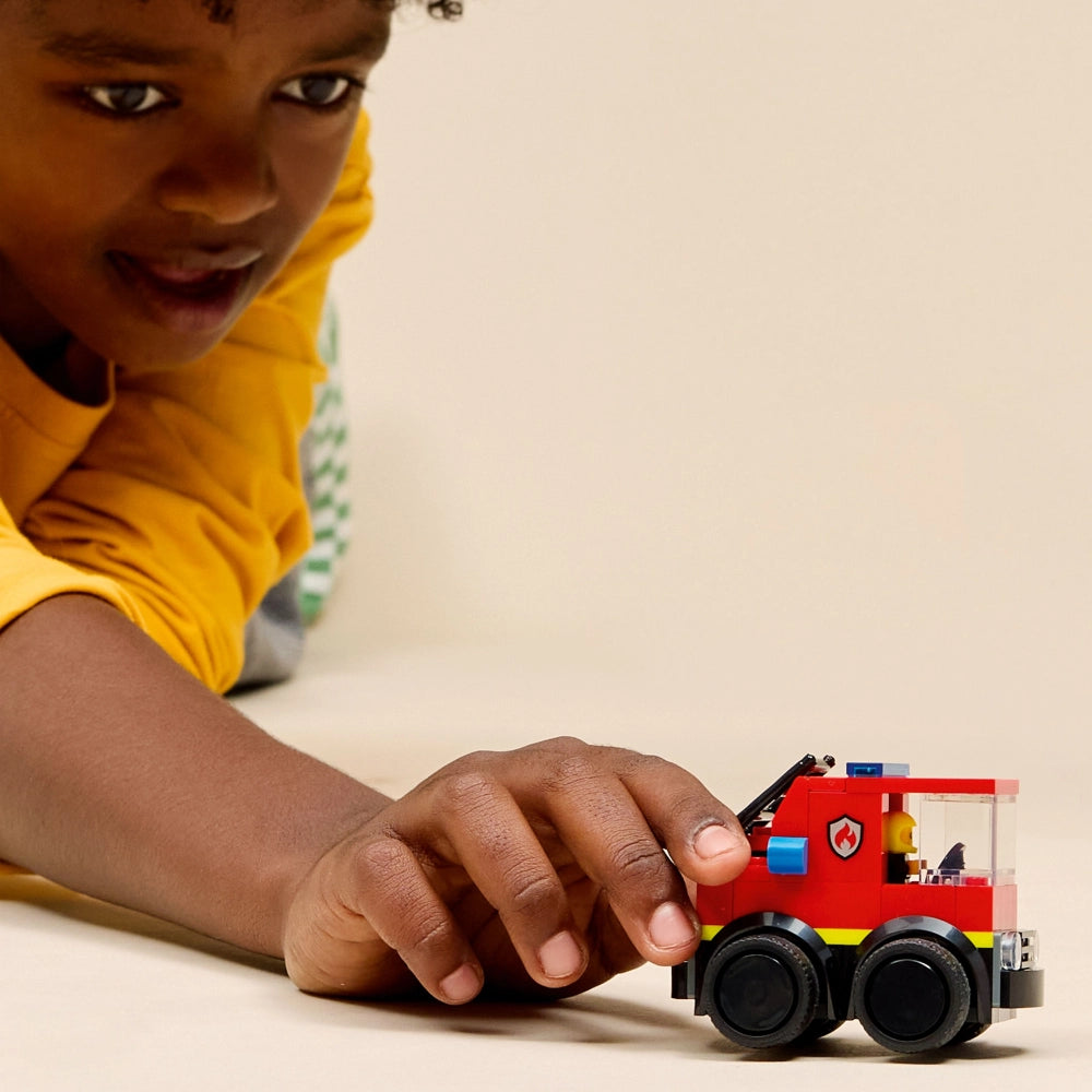 Child in yellow shirt playing with red LEGO fire truck toy featuring black wheels and blue and yellow details on white surface