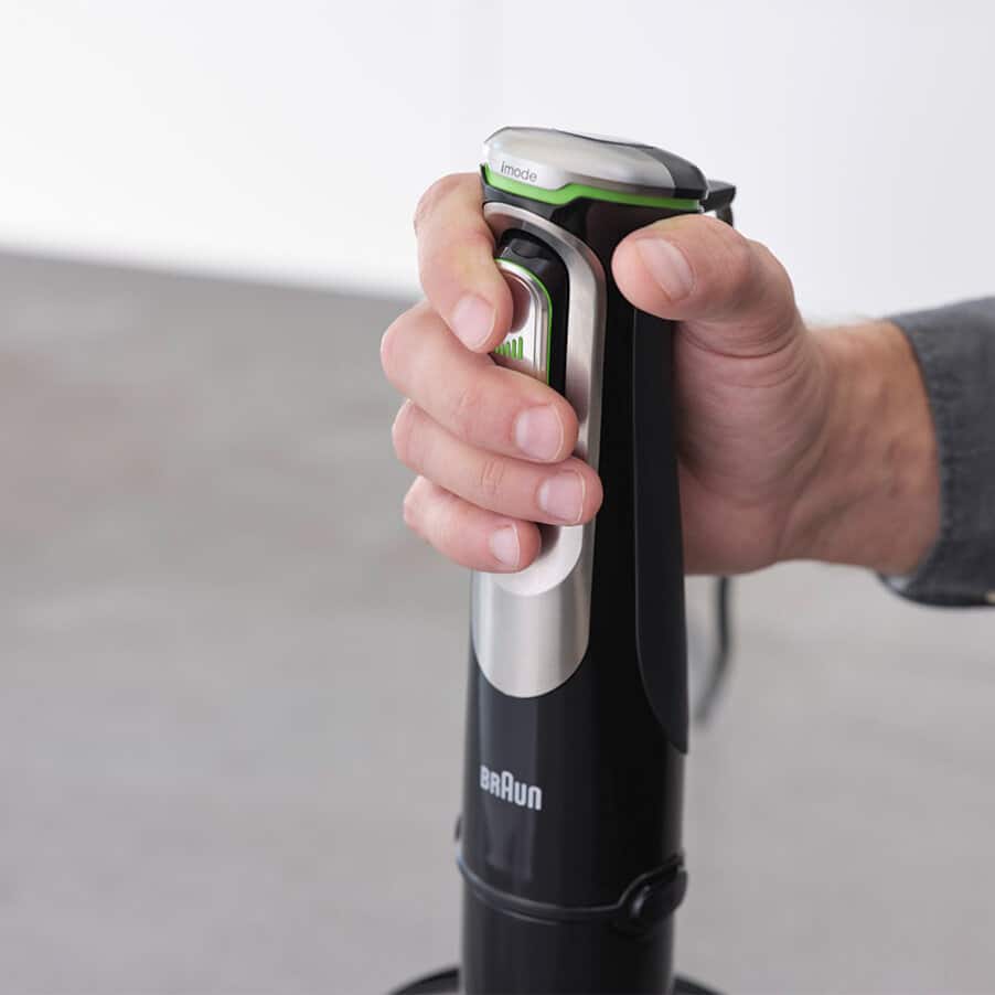 Hand holding a black and silver Braun MultiQuick 9 hand blender with green accent stripe and mode indicator on top, displaying the product's ergonomic design against a neutral background