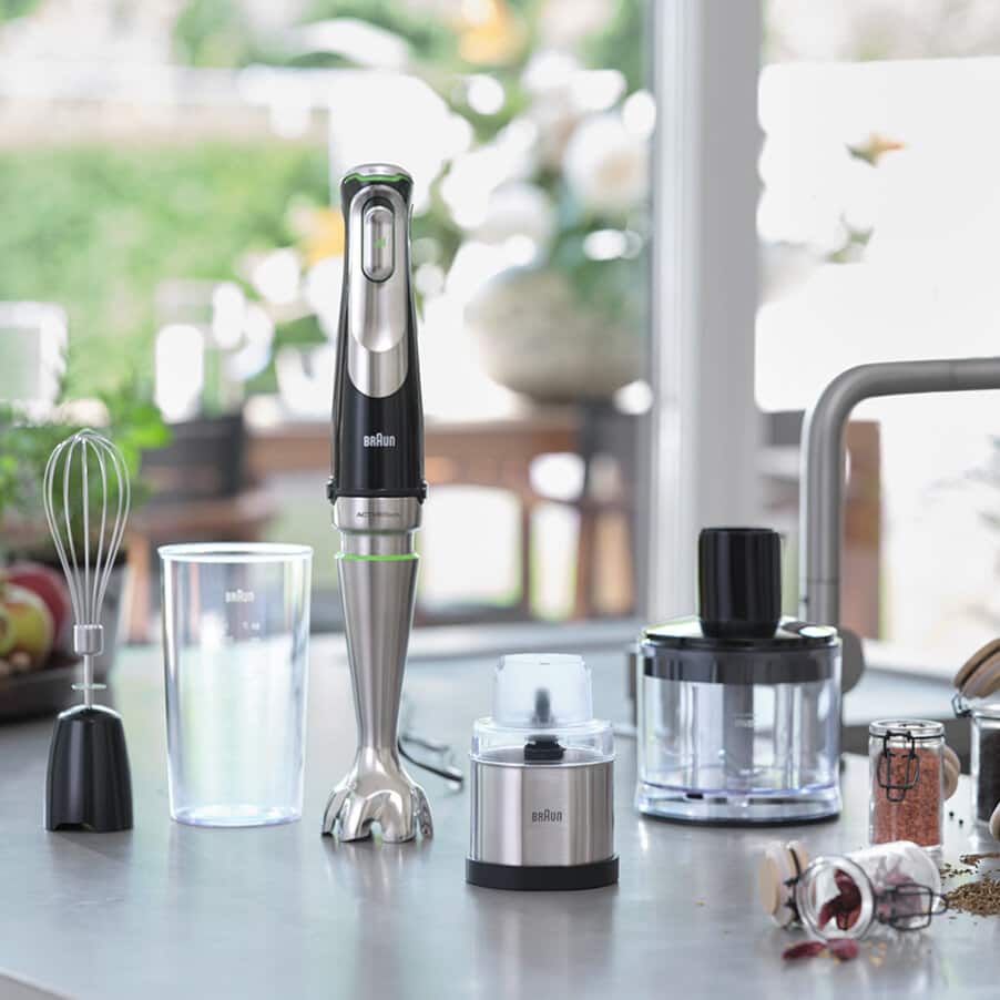 Braun MultiQuick 9 hand blender with stainless steel shaft and black top displayed on kitchen counter with five attachments including whisk, chopper bowl, and measuring cup, surrounded by fresh ingredients and flowers in bright window setting