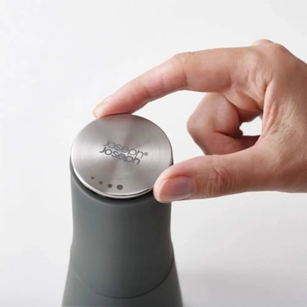 Hand holding a Joseph Joseph Milltop salt and pepper set with stainless steel top and grey cylindrical base against white background