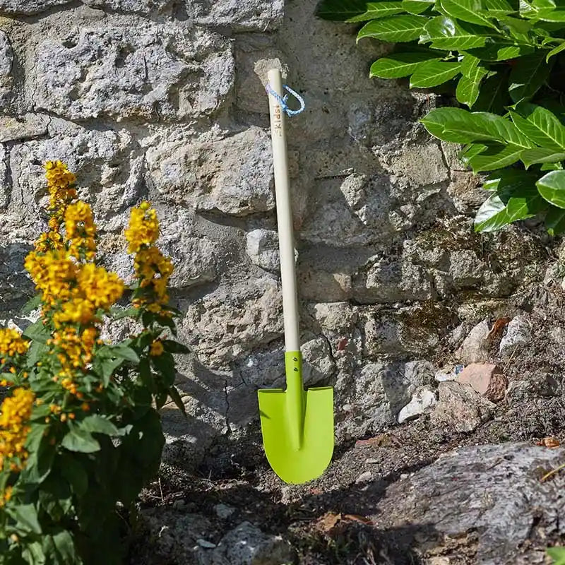 Bright lime green children's garden spade with white handle leaning against weathered stone wall, surrounded by yellow flowering plants and green foliage