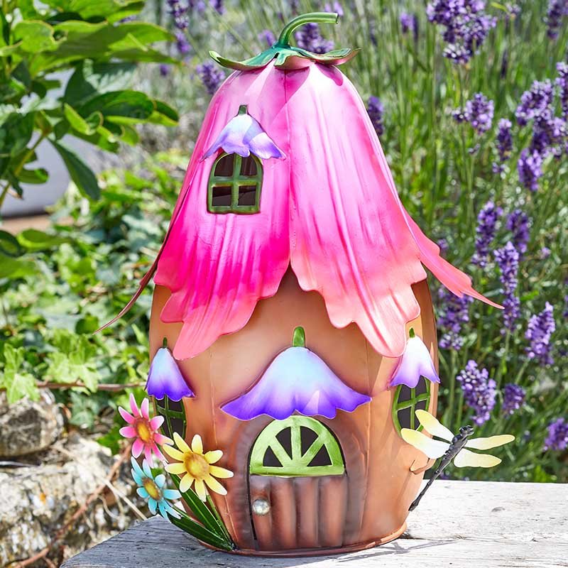 Whimsical flower-shaped garden ornament with pink fuchsia petals forming the roof, tan ceramic body, purple bell-shaped flowers on sides, decorative window and door details, green stem top, surrounded by lavender plants and garden flowers