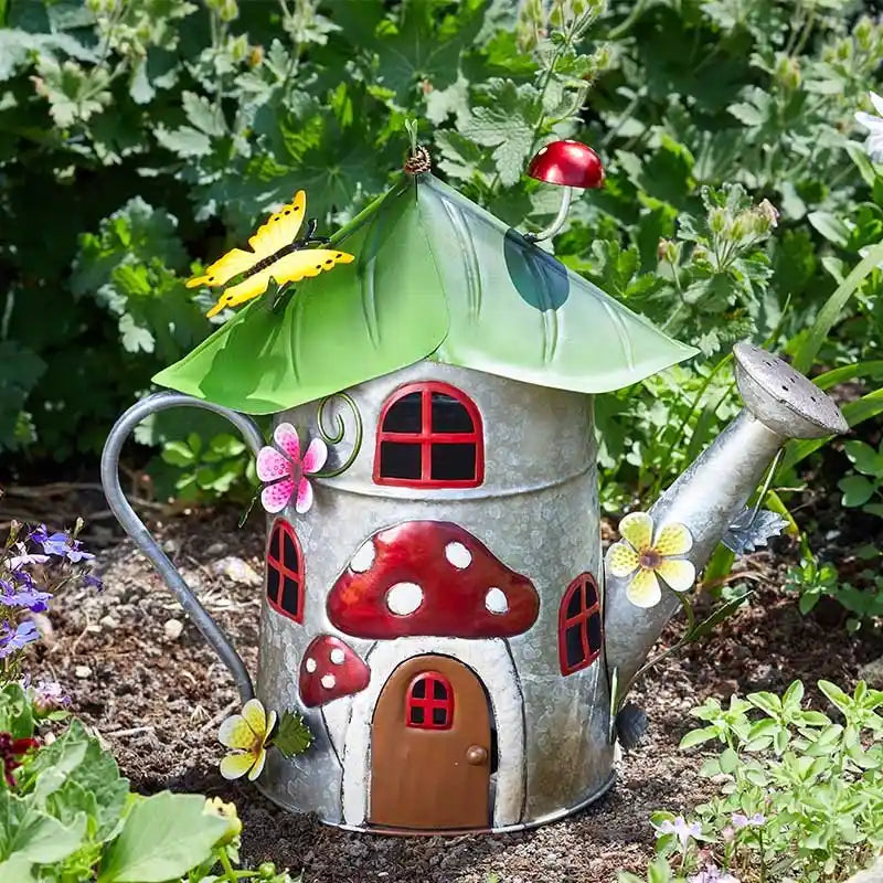 Whimsical mushroom-shaped garden watering can ornament with red and white spotted roof, metal construction, decorative windows and door, surrounded by green foliage and yellow flowers in a garden setting