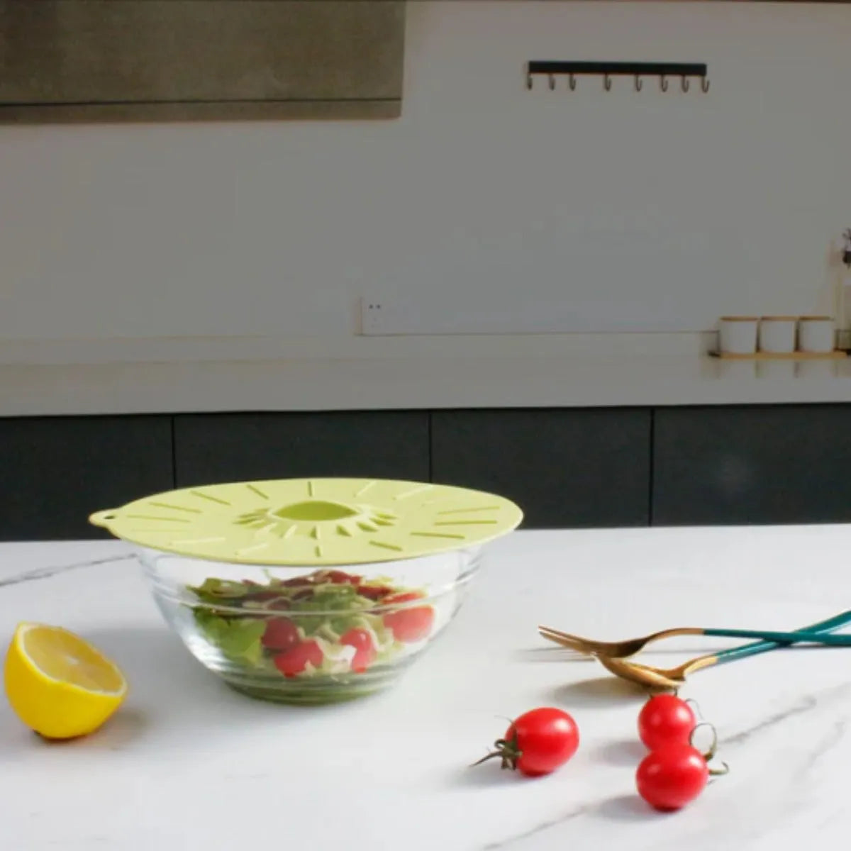 Yellow silicone suction lid covering a glass bowl of salad in a modern kitchen, with fresh tomatoes, lemon half, and kitchen shears on white countertop