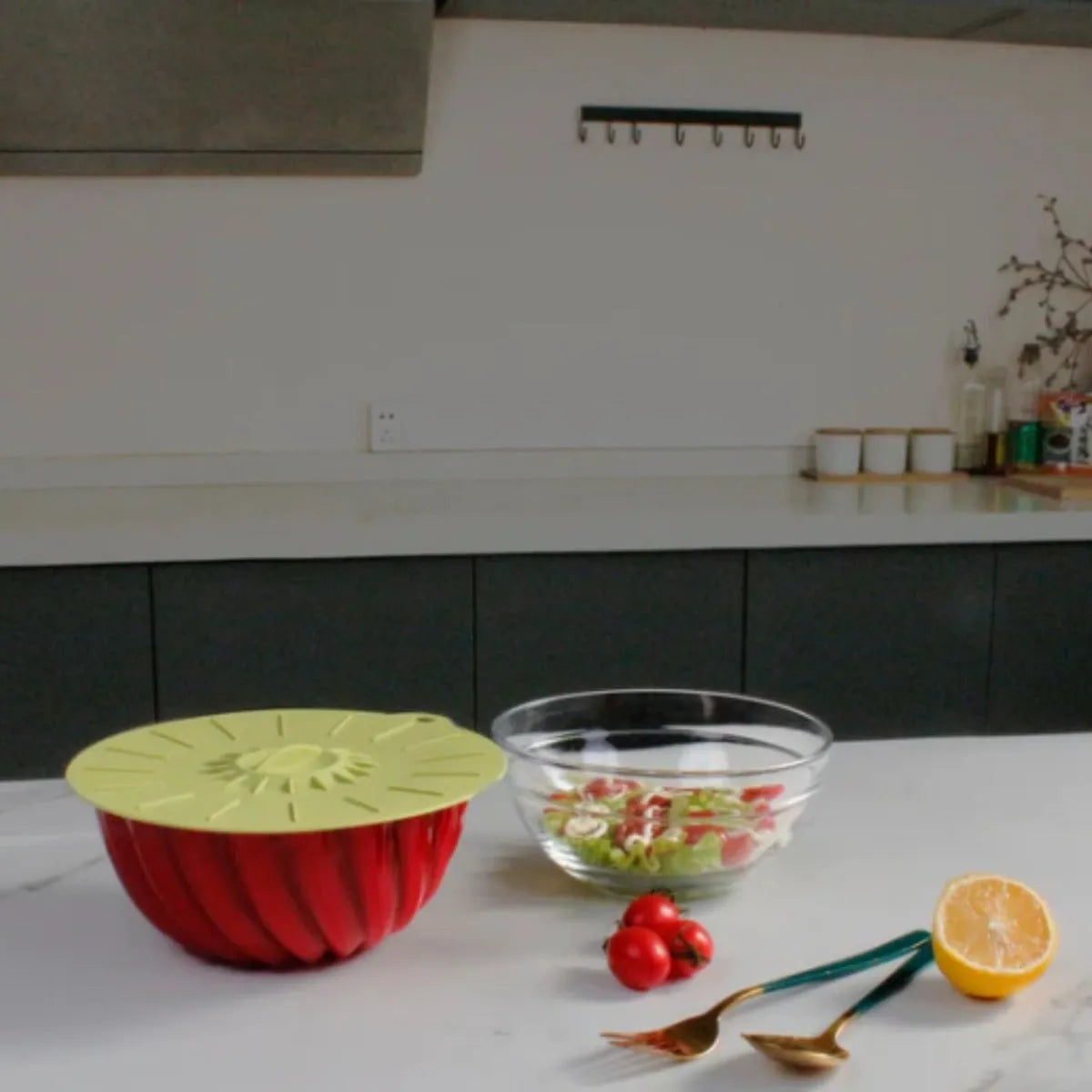 Red silicone bowl with yellow suction lid next to glass bowl containing salad, with tomatoes, lemon half and gold serving utensils on white kitchen counter