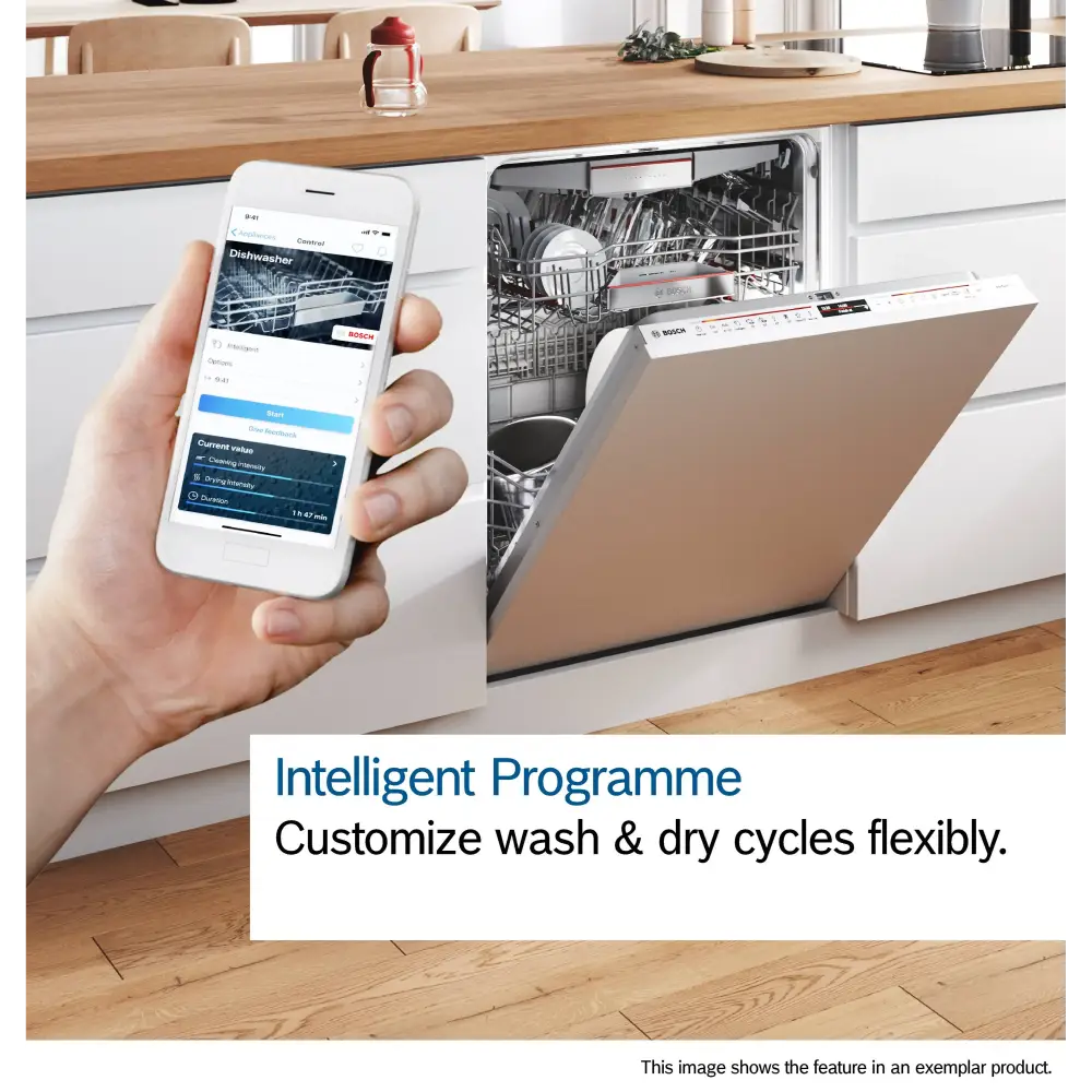White hand holding smartphone displaying Bosch dishwasher app with wash cycle controls, positioned next to integrated Bosch Series 6 dishwasher with beige door panel in modern white kitchen with wooden countertop, demonstrating intelligent programme customization feature