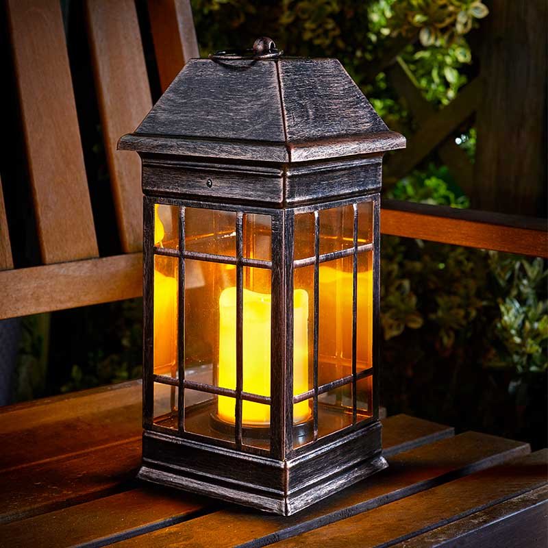 Smart Solar Seville solar lantern with dark metal frame and amber glass panels emitting warm yellow light, positioned on wooden garden furniture at night with foliage visible in background