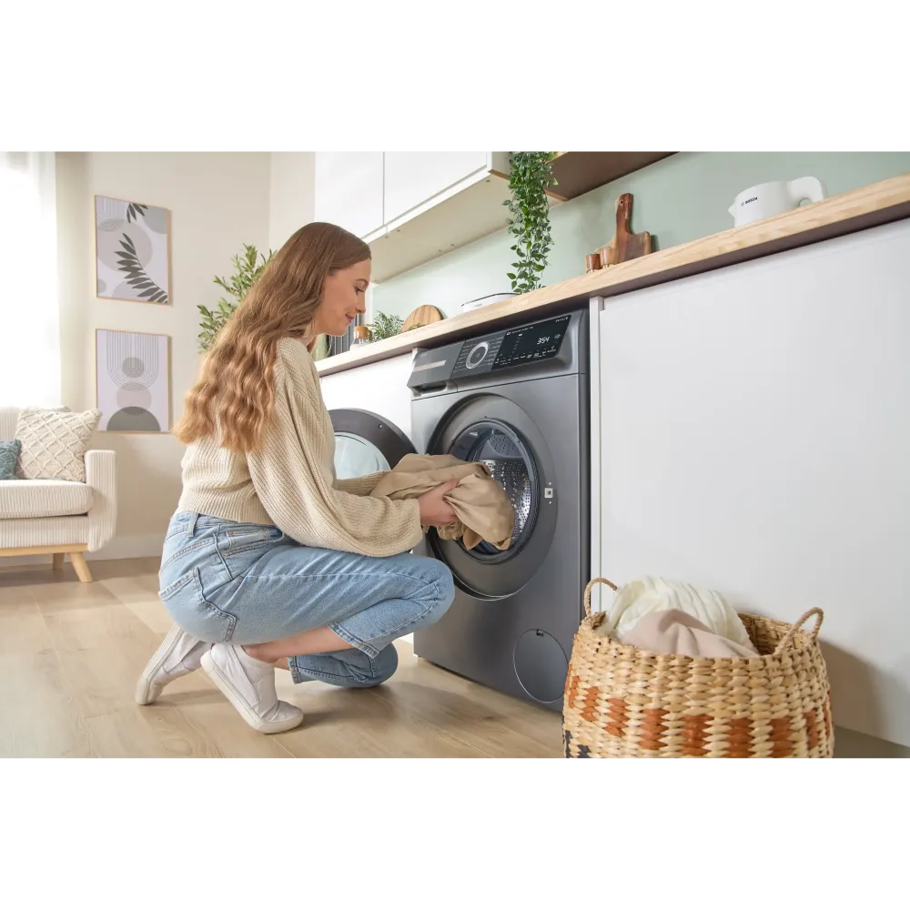 Woman loading laundry into a dark grey Bosch Series 6 washing machine in a modern utility room with white cabinetry, wooden shelving, potted plants, and a wicker laundry basket