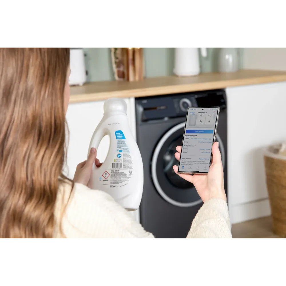 Woman holding smartphone displaying washing machine app controls next to grey Bosch Series 6 front-load washing machine in modern white and wood kitchen with detergent bottle