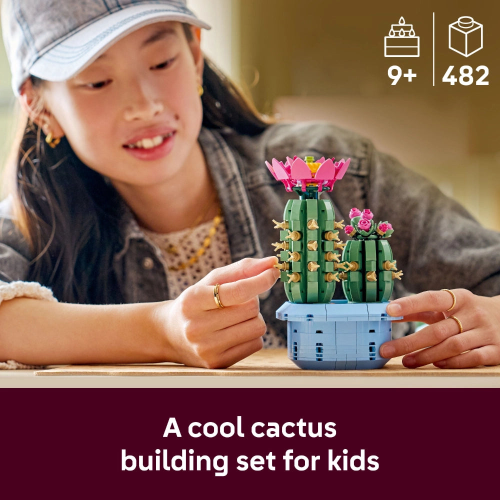 A young girl wearing a gray cap examines a LEGO Flowering Cactus building set featuring green segmented cactus pieces with pink flowers and a light blue pot, positioned on a wooden surface
