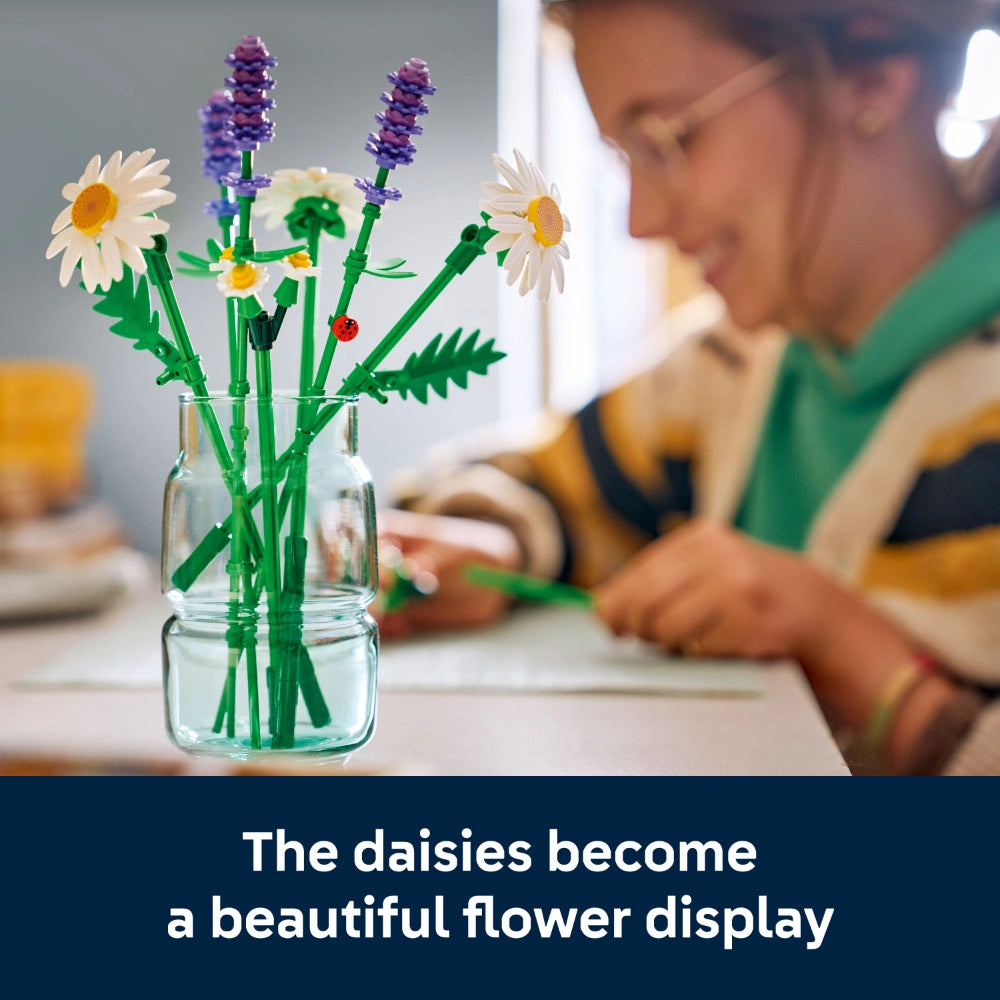 LEGO Daisies flower building set displayed in a clear glass vase with water, featuring white daisy blooms with yellow centers, purple lavender flowers, and green stems, with a child in a green shirt constructing the set in the blurred background