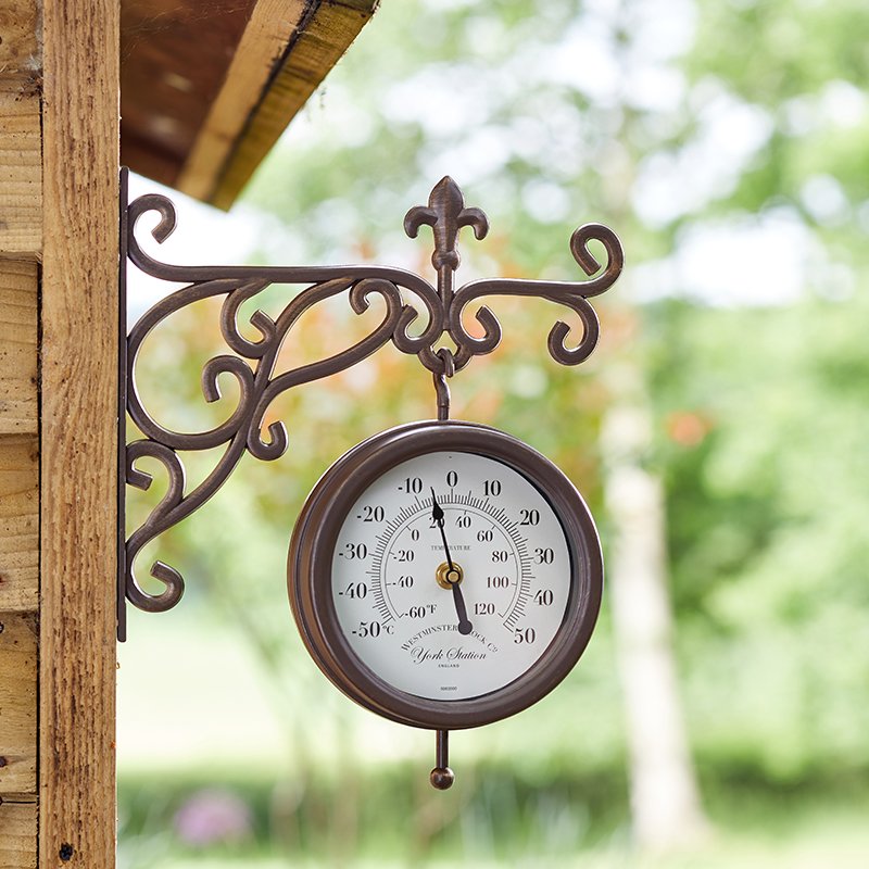 Vintage-style dark bronze metal garden clock and thermometer mounted on wooden wall, featuring ornate scrollwork bracket and white dial face displaying temperature and time scales, with blurred green garden foliage in background
