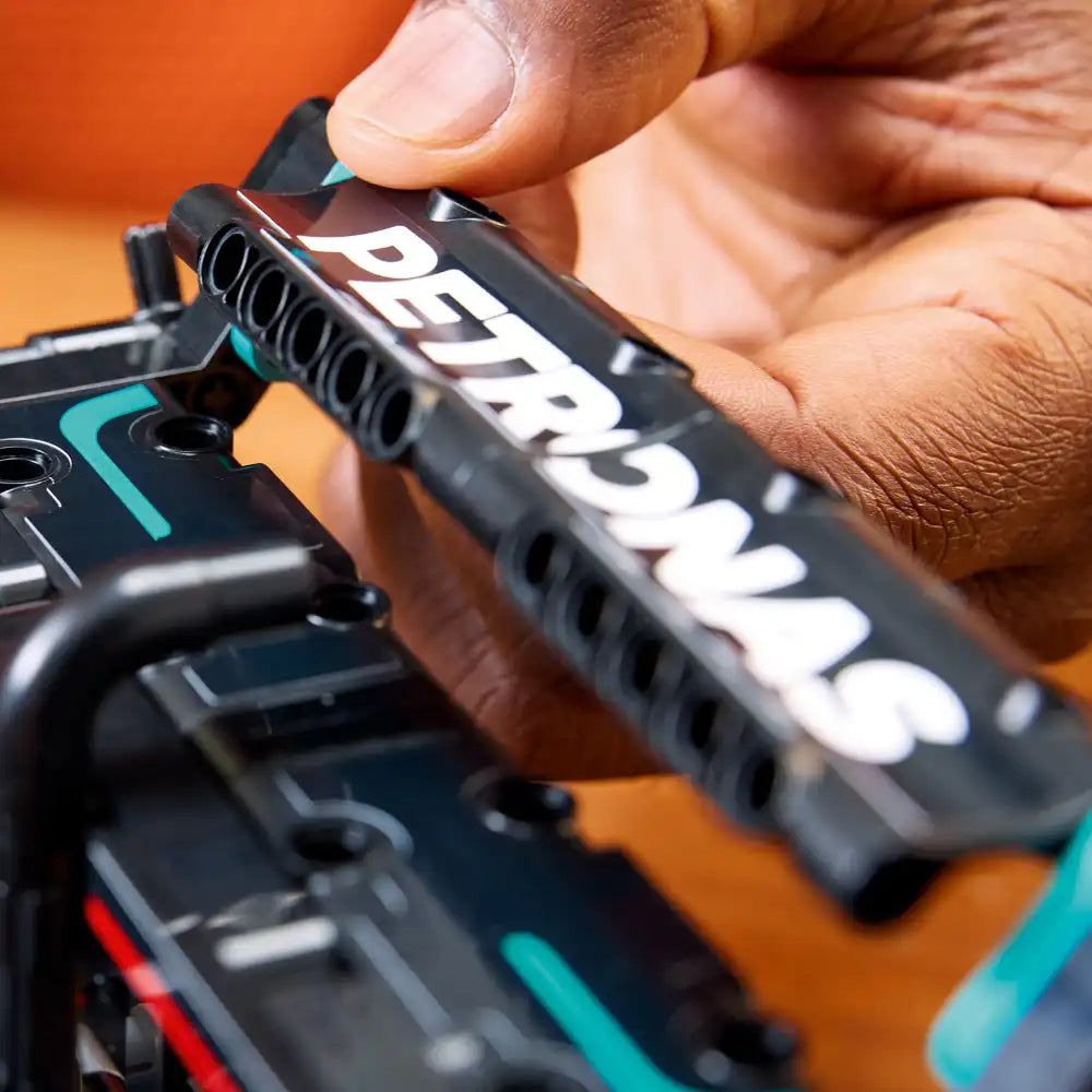 Close-up of hands holding black LEGO Technic Mercedes-AMG F1 W14 E Performance model 42171 showing white text on dark plastic pieces with turquoise and red accent details against orange background