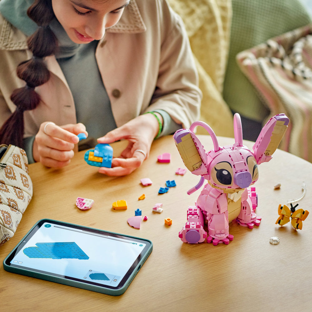 Young child building pink LEGO Angel figure at wooden table with scattered colorful bricks, tablet displaying building instructions nearby, wearing beige cardigan over green shirt