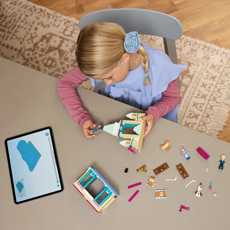 Child with blonde hair in braids wearing a blue and pink long-sleeved shirt sitting at a table with LEGO Frozen Arendelle Castle set 43265, displaying colorful castle pieces, minifigures and accessories alongside an iPad showing the building instructions