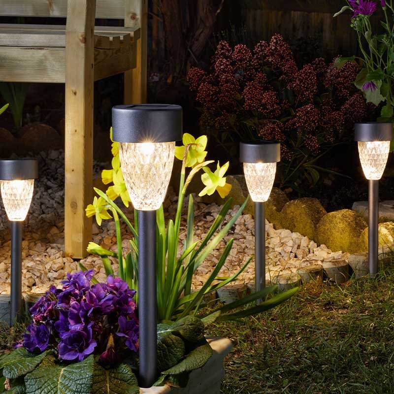 Four solar garden stake lights with textured glass shades and dark metal stems illuminating a garden bed at night, surrounded by purple petunias, yellow flowers, burgundy seed heads, ornamental grasses, and decorative gravel near wooden decking