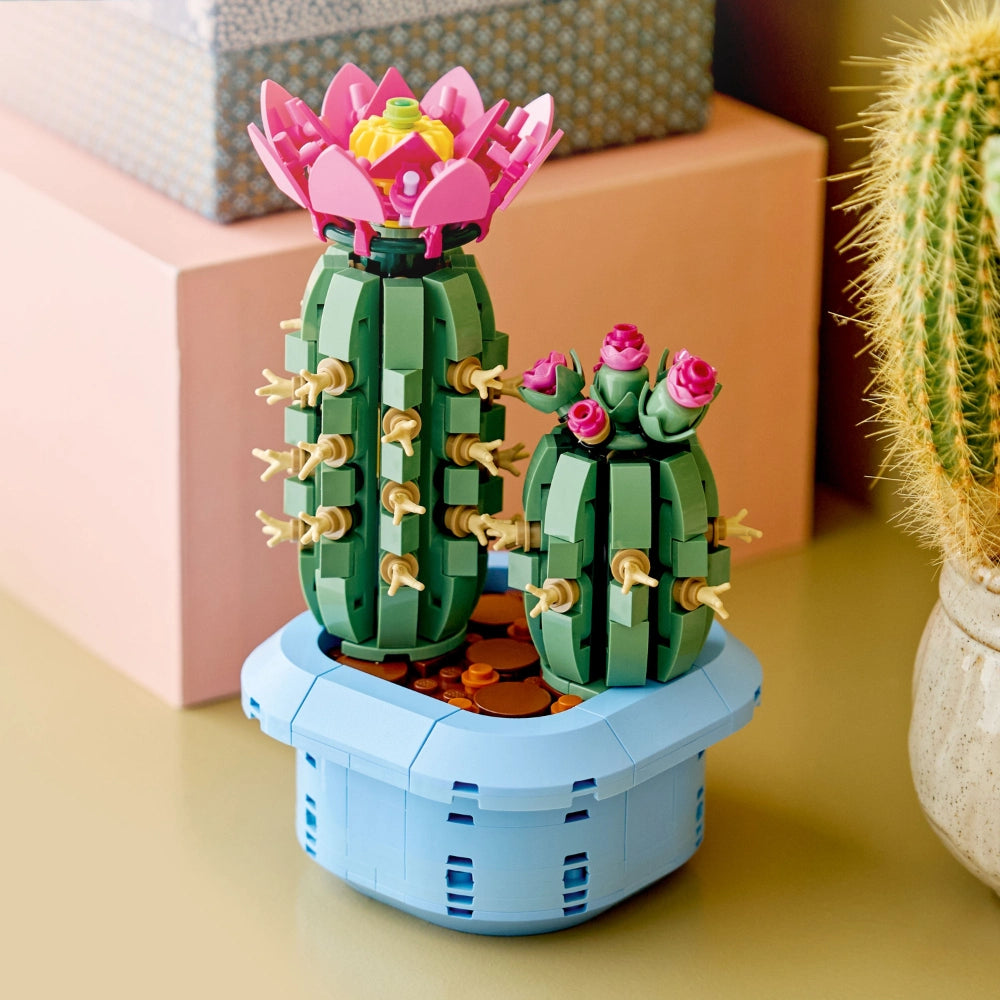 LEGO Flowering Cactus set 11509 featuring two green segmented cacti with gold spines in a light blue pot, topped with pink and magenta blooms and yellow centers, displayed on a yellow surface alongside decorative boxes and real potted plants