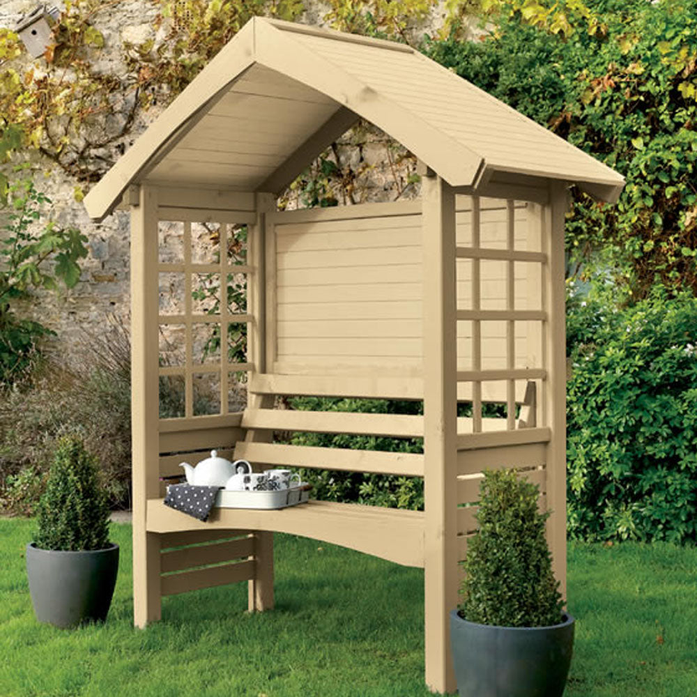 Cream-coloured wooden garden arbour with peaked roof, lattice windows and built-in bench seat in lawn setting with ivy and potted topiary plants