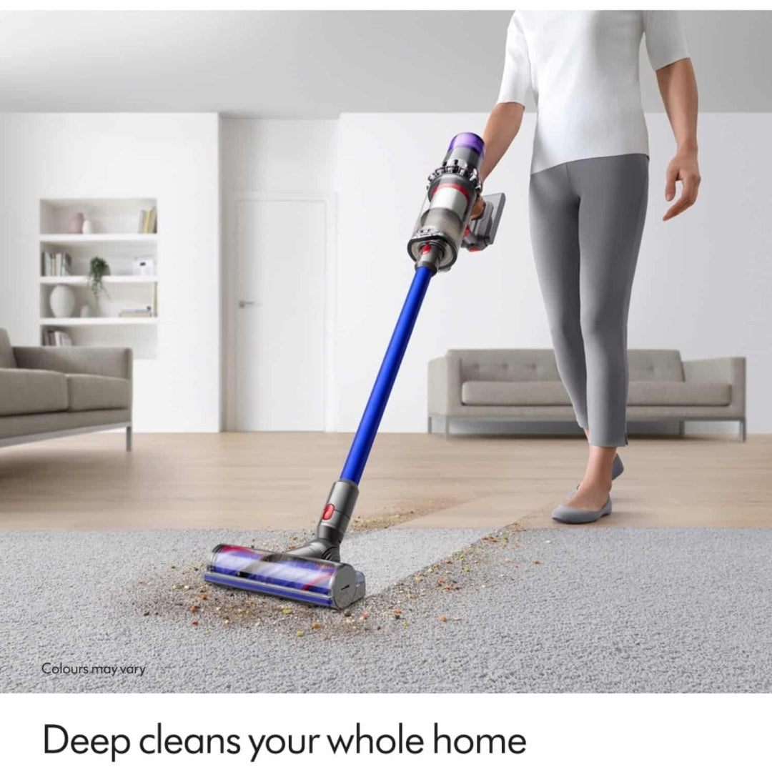Woman using blue and purple Dyson V11 cordless vacuum with motorized brush head to clean light gray carpet in modern minimalist living room with white shelving and gray sofa