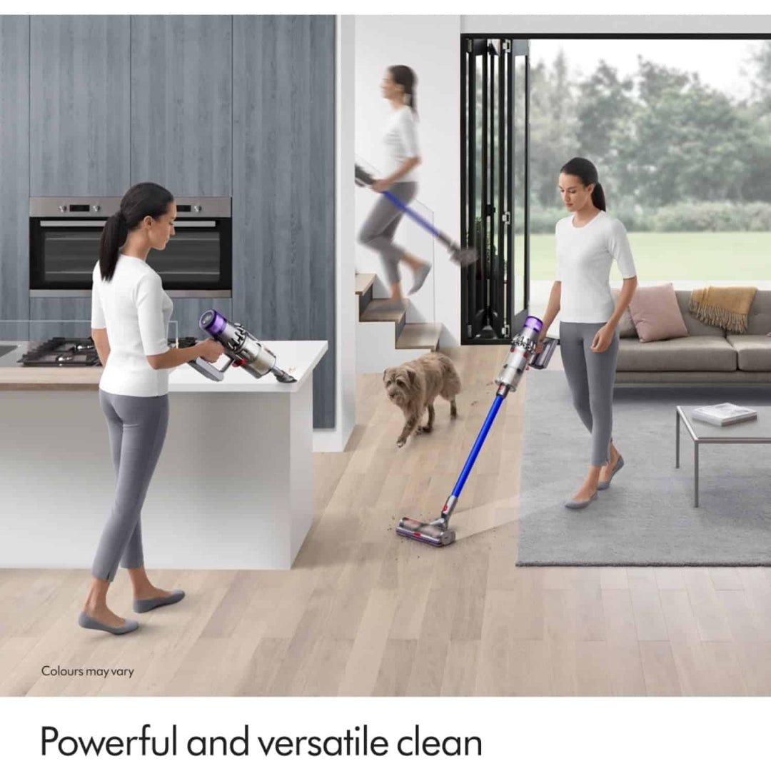 Woman using Dyson V11 cordless vacuum cleaner with purple and blue design across modern kitchen, hallway and living room spaces, demonstrating versatile cleaning on wooden floors and carpets with dog nearby