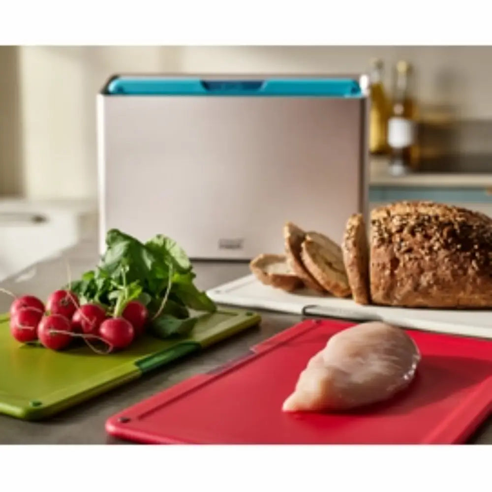 Joseph Joseph Folio Icon 4 piece chopping board set in multicolour featuring a lime green board with fresh radishes, red board with raw chicken breast, and sliced brown bread on white board, with turquoise storage case in background on kitchen counter