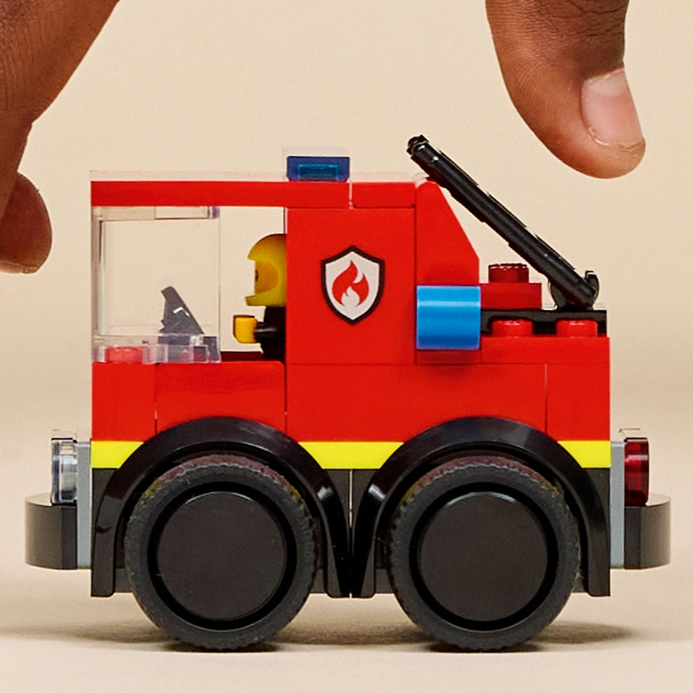 LEGO fire truck set 60482 featuring a red and yellow brick-built emergency vehicle with black wheels, blue light, firefighter minifigure, and articulated ladder, displayed against a cream background with child's hand for scale